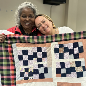 image of two women smiling and holding up a colorful patchwork quilt at Gee's Bend
